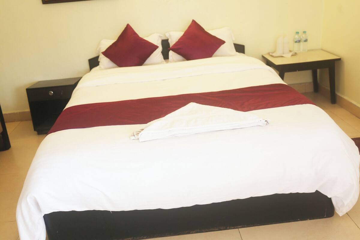 A neatly made bed with white and red bedding, a folded towel, and a nightstand with a lamp and water bottles.