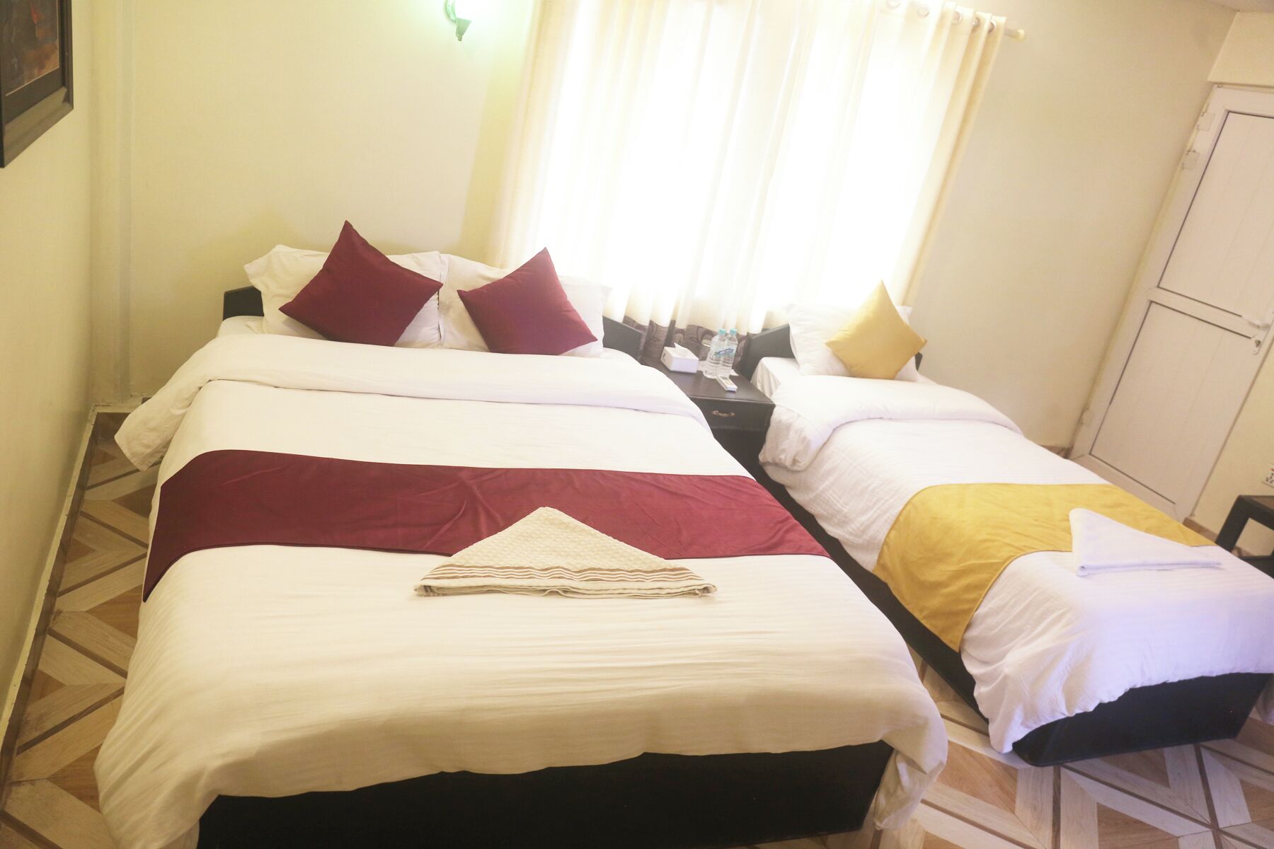 Een hotelkamer met twee bedden, elk met witte en bordeauxrode beddengoed, een gestreept handdoek, en een nachtkastje met een lamp en een fles.
