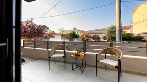 Een balkon met twee rieten stoelen en een kleine tafel met drankjes, met uitzicht op een woonwijk met huizen en bloeiende bomen.