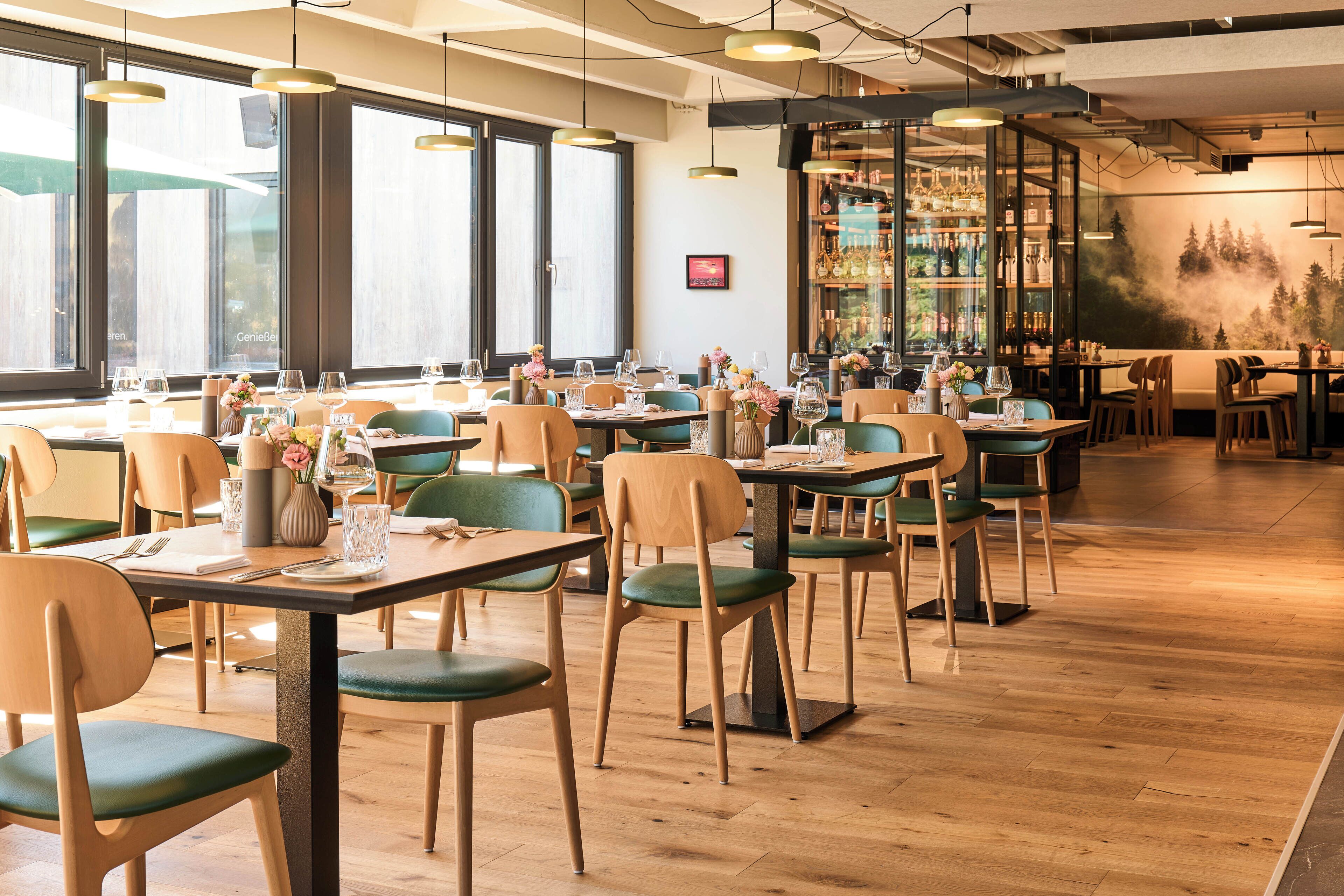 A restaurant with wooden tables and chairs, large windows, and a bar area with shelves of bottles.