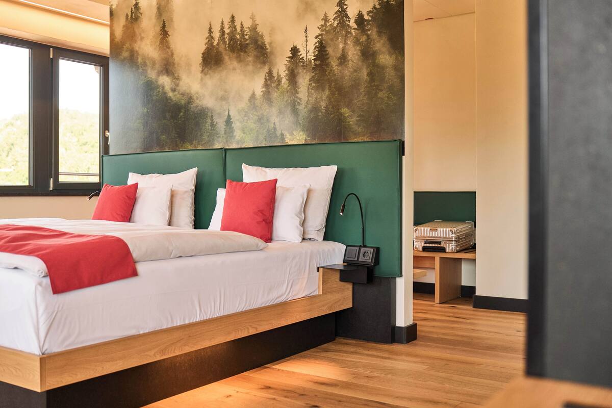 A modern bedroom with a large bed, green headboard, and a forest mural on the wall.