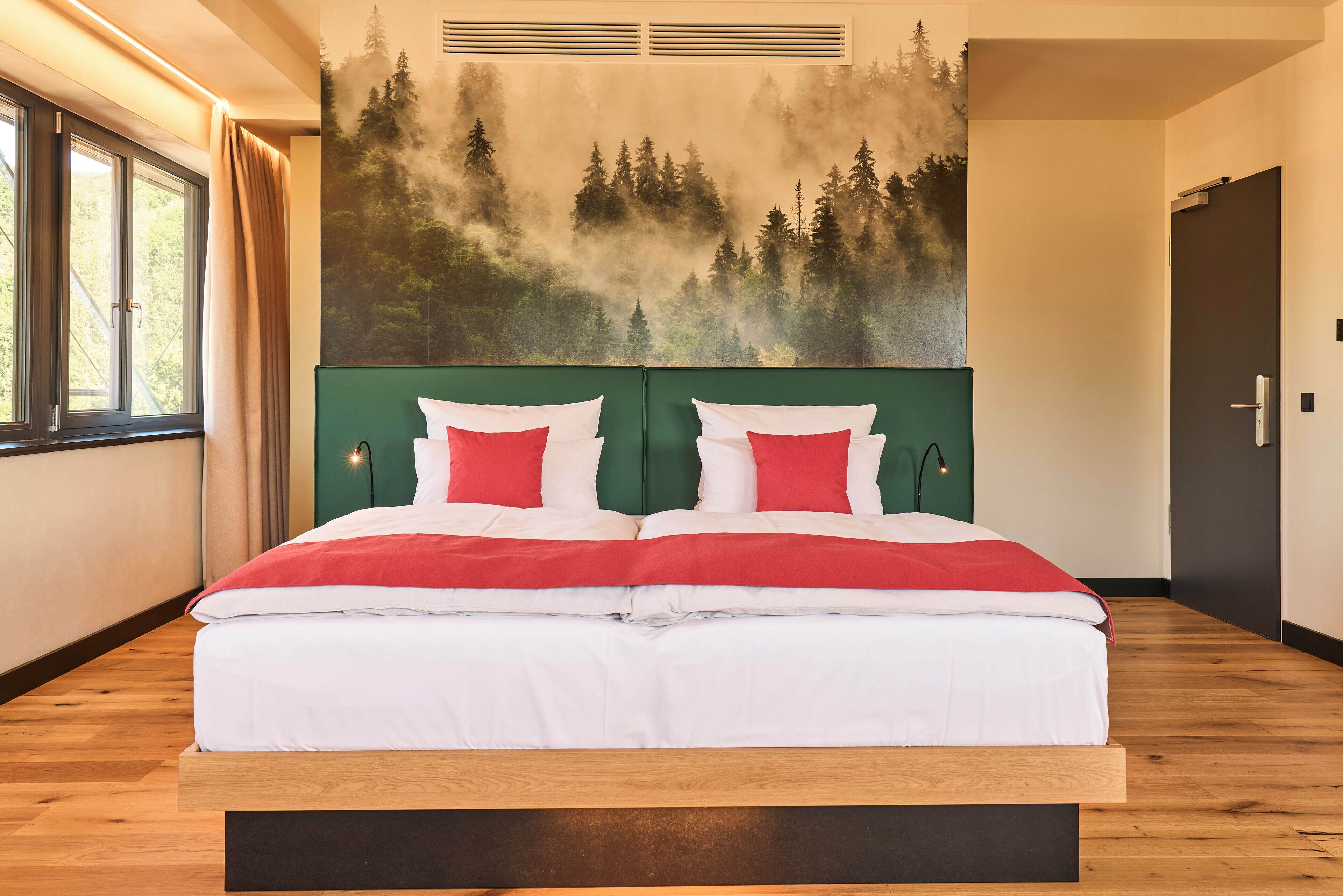 A double bed with white and red bedding, a green headboard, and a forest mural on the wall.
