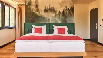 A double bed with white and red bedding, a green headboard, and a forest mural on the wall.