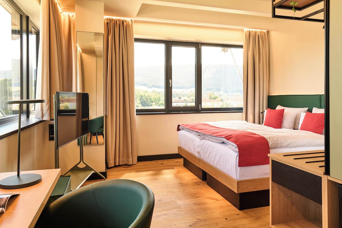 A hotel room with a bed, a desk, a chair, a TV, and large windows with curtains.