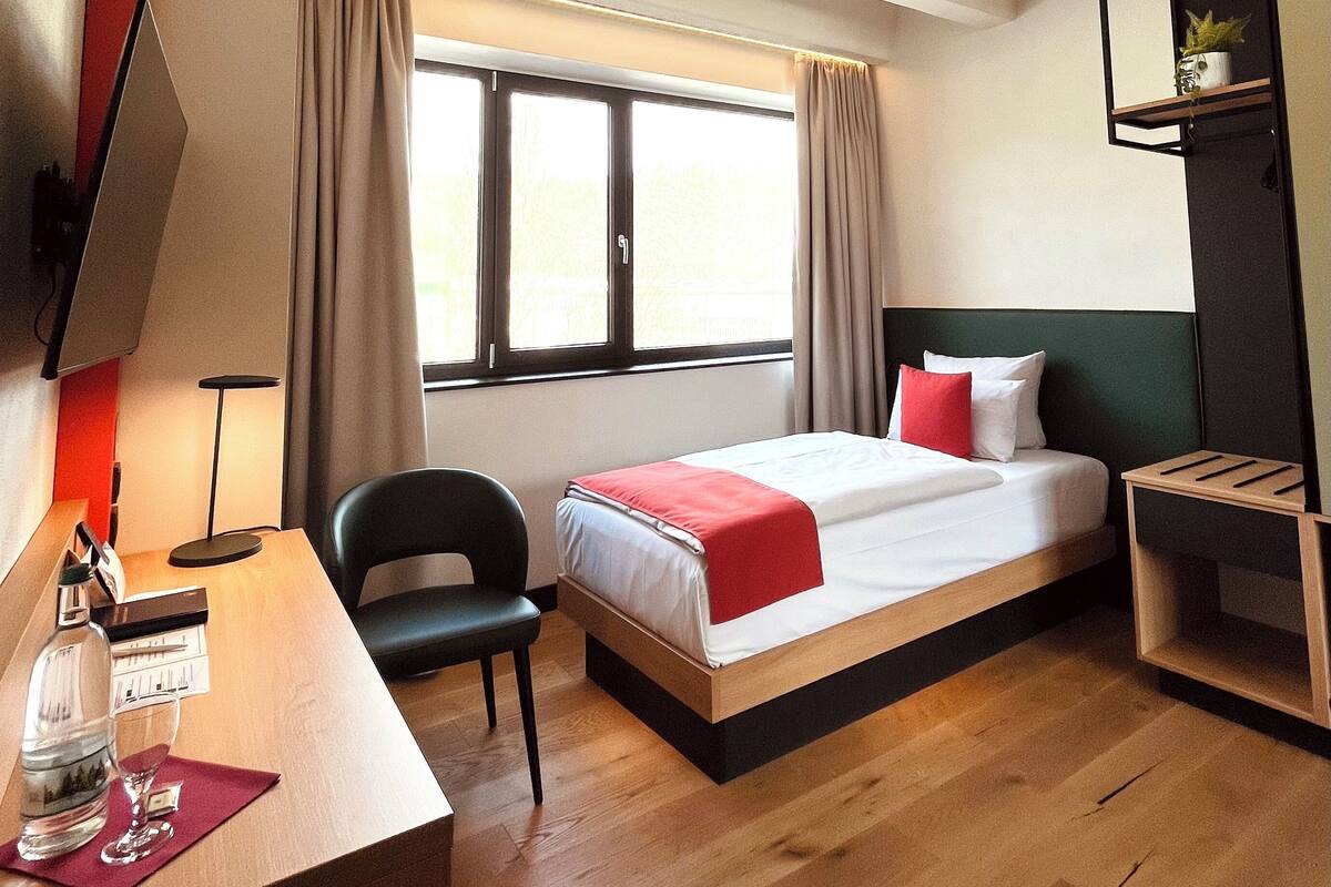 A hotel room with a bed, desk, chair, and a small shelf with a plant.
