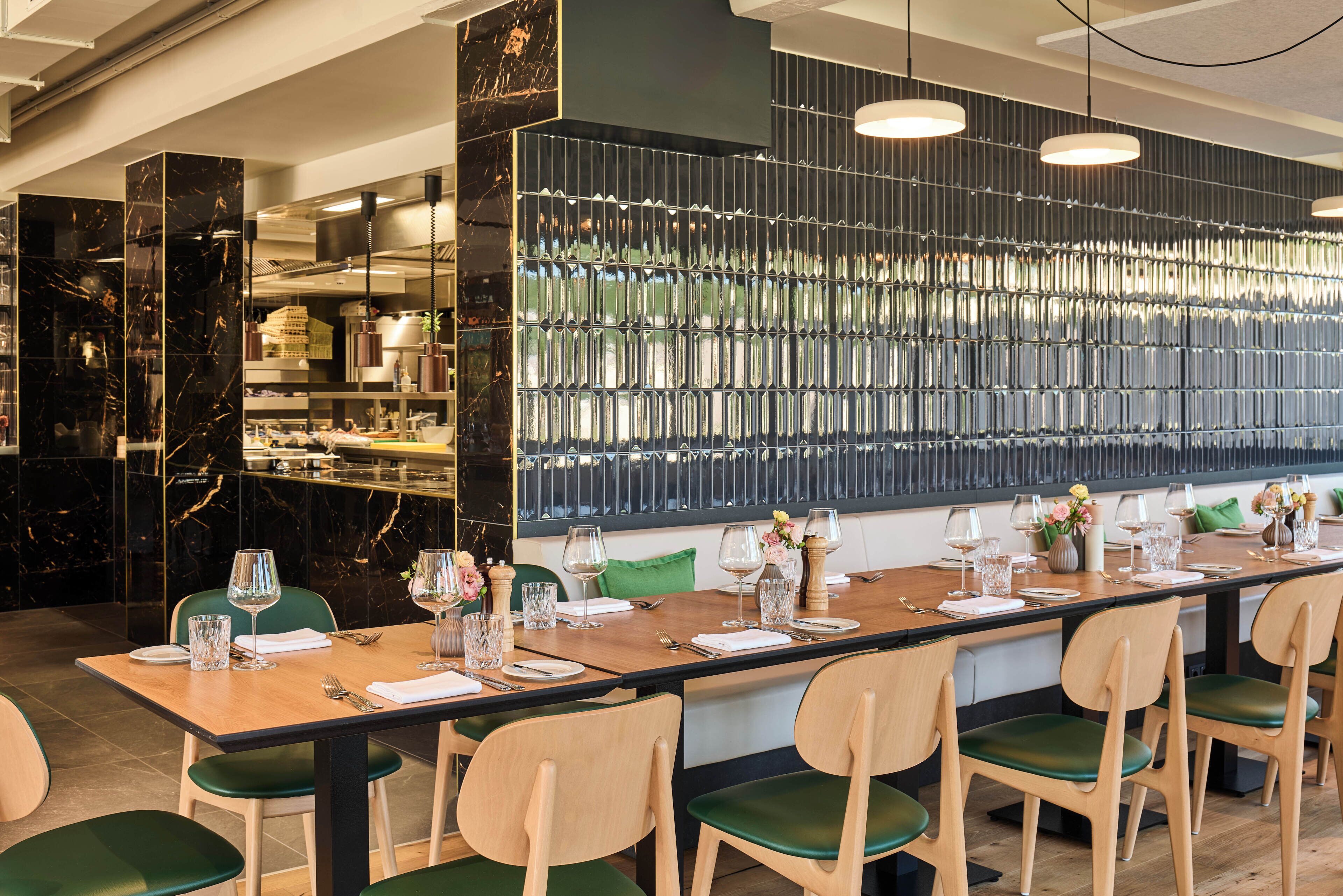 A restaurant with a long wooden table set for dining, green chairs, and a decorative wall with vertical mirrors.