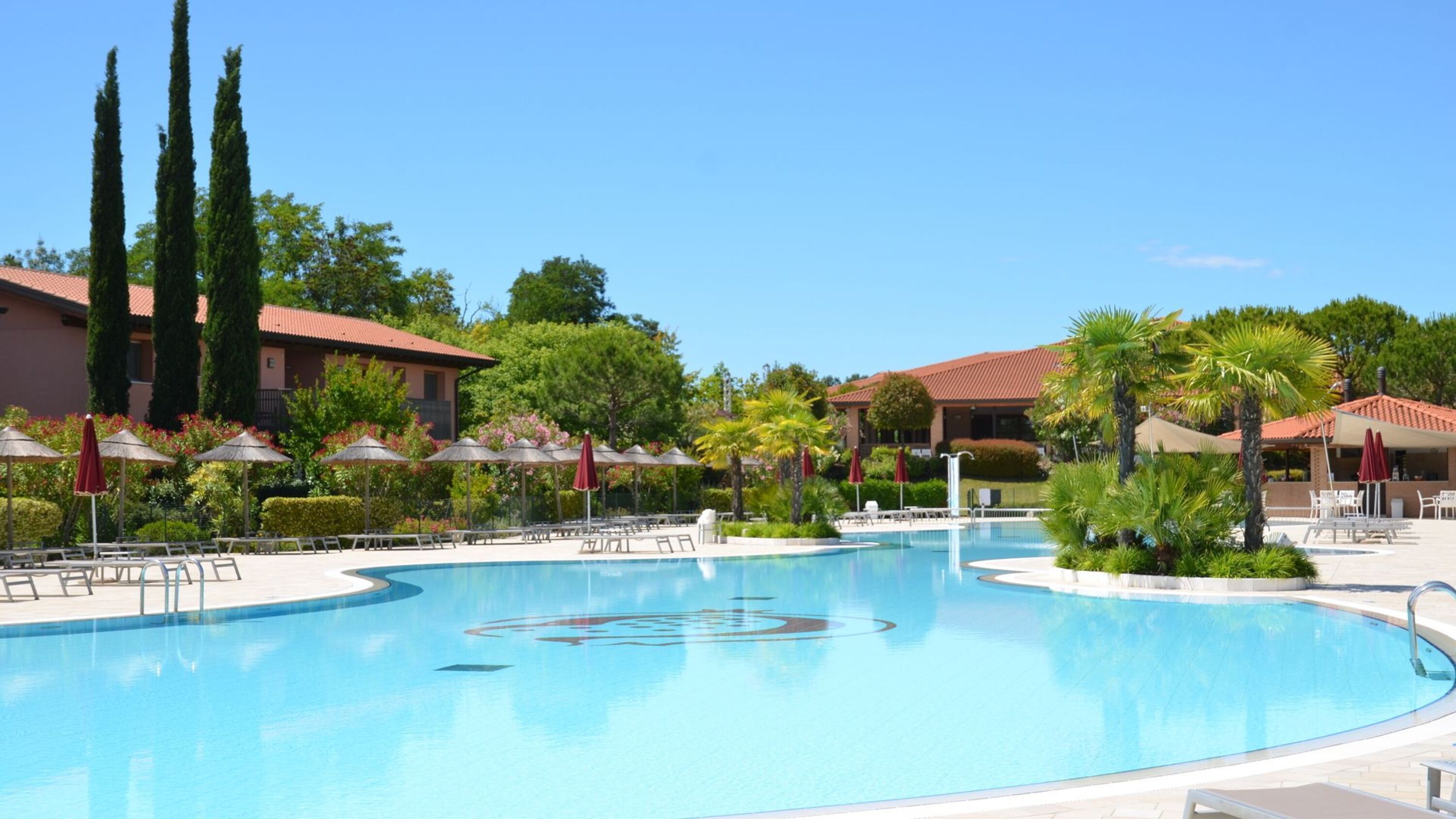A large swimming pool with lounge chairs and umbrellas, surrounded by palm trees and a building with a red roof.