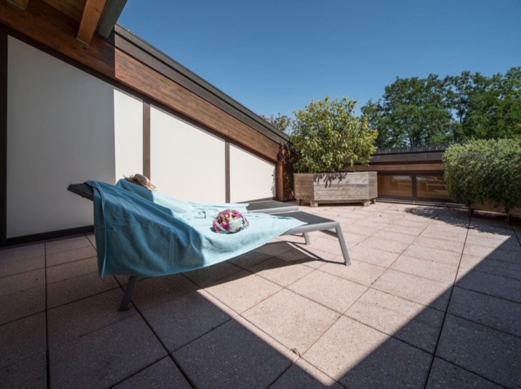 A sunbed in a covered outdoor area with a tiled floor, a wooden pergola, and a stone planter with a tree.