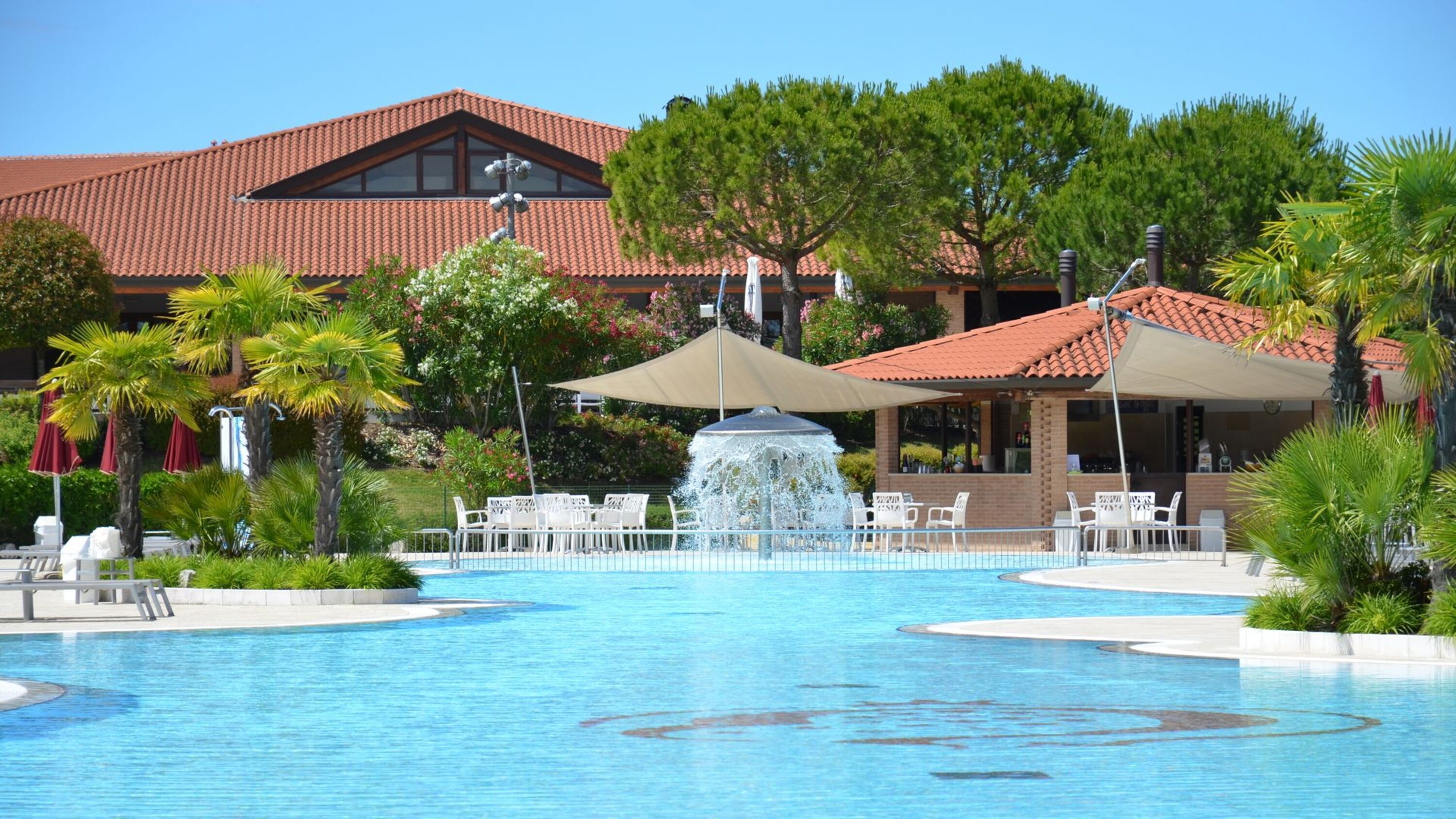 Une piscine avec une fontaine, entourée de palmiers et de chaises longues, avec un bâtiment en arrière-plan.