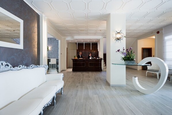 Una moderna hall d'albergo con una reception, un divano bianco e un originale tavolo in vetro con una grande lettera 'G' bianca.