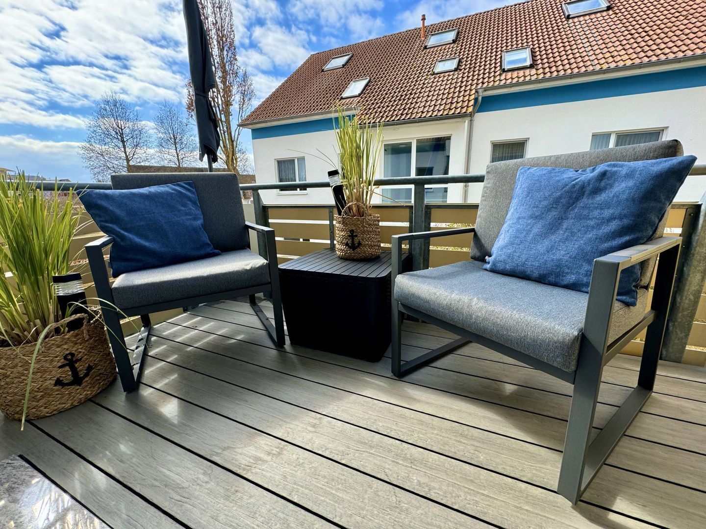 A wooden deck with two grey chairs and a small table, a woven basket with an anchor, and a house with a red-tiled roof in the background.