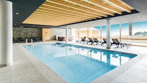 A modern indoor swimming pool with clear blue water, surrounded by lounge chairs and a stone wall.