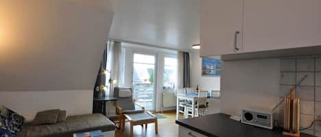 Ein modernes Wohnzimmer mit einer Couch, einem Esstisch mit Stühlen, einer Küchenzeile mit Schränken und einem Radio sowie einem großen Fenster mit Vorhängen.