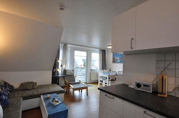 Ein modernes Wohnzimmer mit einer Couch, einem Esstisch mit Stühlen, einer Küchenzeile mit Schränken und einem Radio sowie einem großen Fenster mit Vorhängen.