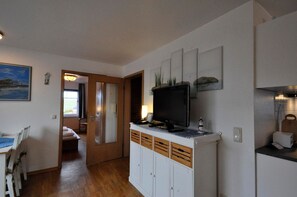 A room with a television on a cabinet, a dining table with chairs, a door leading to another room, and a painting on the wall.