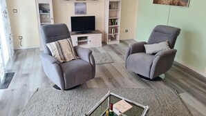 Una sala de estar con dos sillones grises, un televisor de pantalla plana, una estantería y un cuadro con un atardecer.