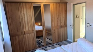 Un dormitorio con un armario grande de madera, una cama con ropa de cama blanca y un escritorio pequeño con una lámpara.