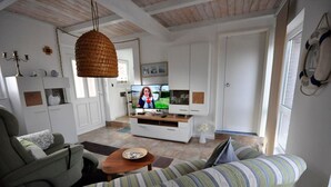Een woonkamer met een bank, een salontafel en een televisie. Er hangt een lichtarmatuur van riet en er staat een witte kast met glazen deuren.