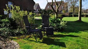Een tuin met twee zwarte stoelen en een tafel, omgeven door groen en een bakstenen gebouw op de achtergrond.