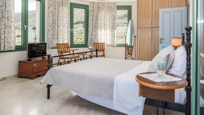Une chambre à coucher avec un lit, des chaises en bois, une commode, une table avec une lampe, et une vue sur la verdure par les fenêtres.