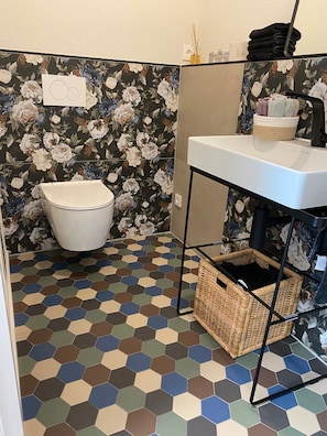 Un baño con un lavabo blanco, un inodoro, un suelo con estampado hexagonal y papel tapiz floral.