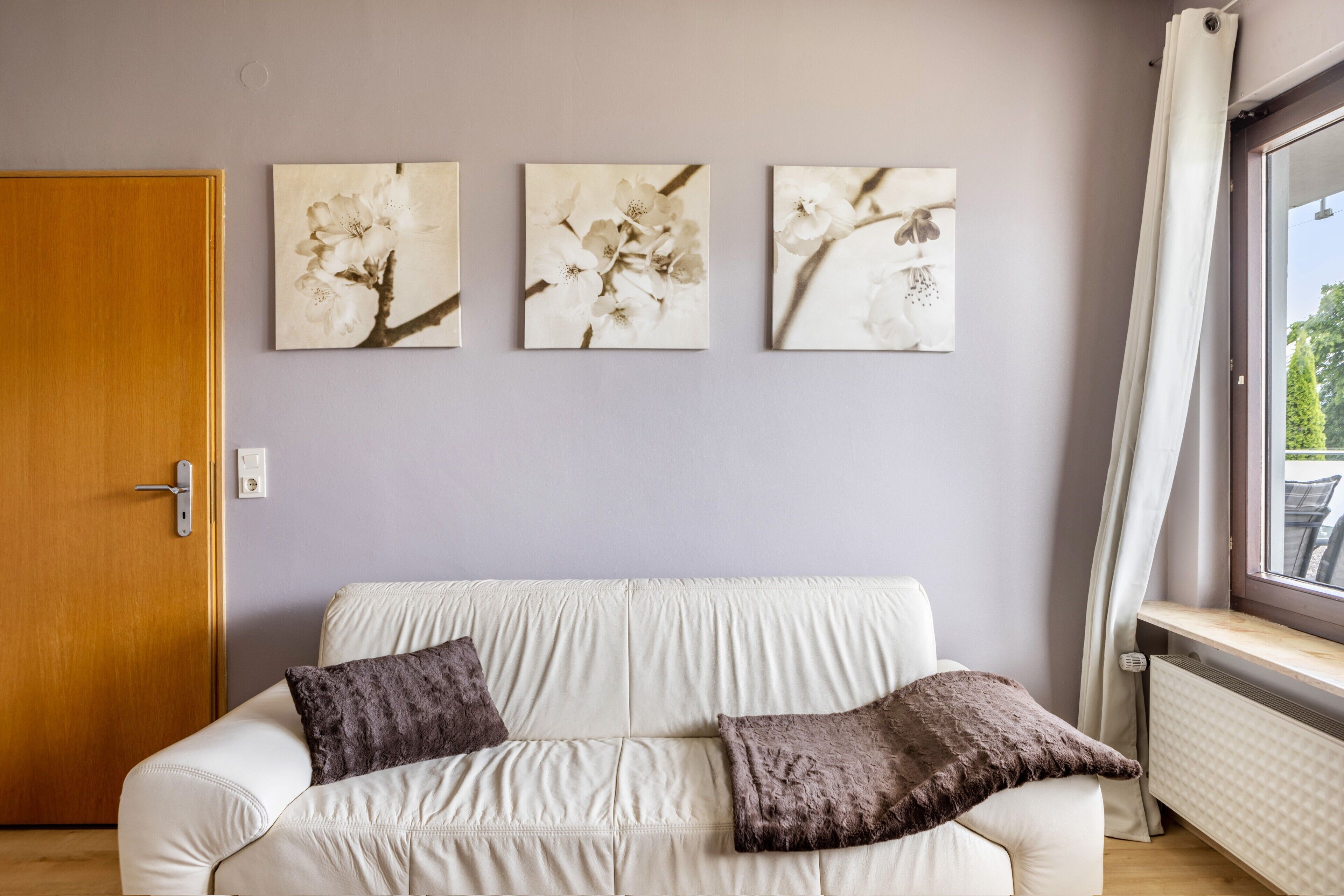 Ett rum med en vit soffa, tre blomstermotiv som hänger på väggen, en trädörr och ett fönster med utsikt.