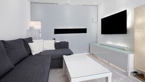 Ein modernes Wohnzimmer mit einem Ecksofa, einem Glastisch, einem Flachbildfernseher an einer weißen Wand und einem schlichten Konsolentisch.
