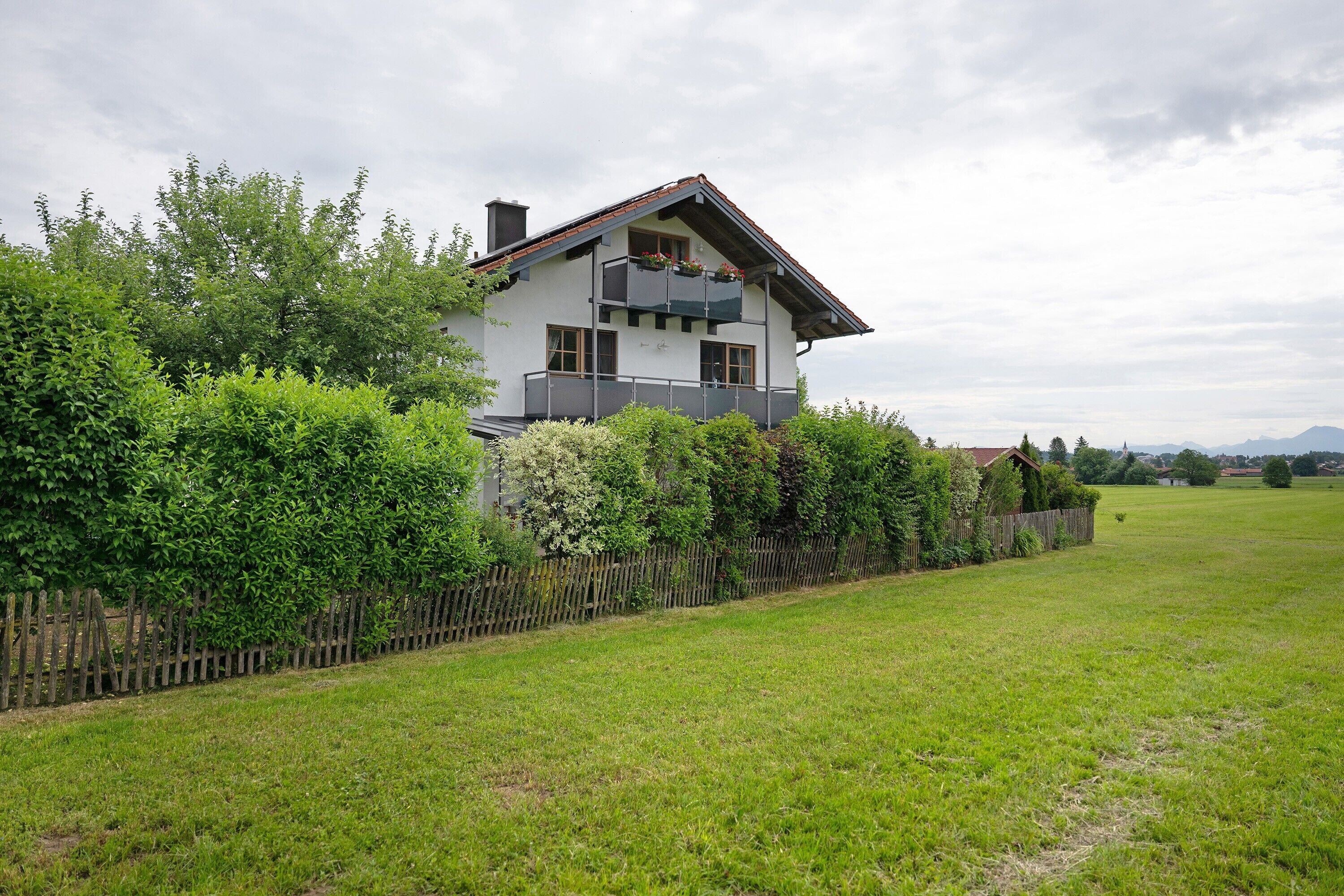 Ein zweistöckiges Haus mit Balkon, umgeben von einem Holzzaun und üppiger Bepflanzung.