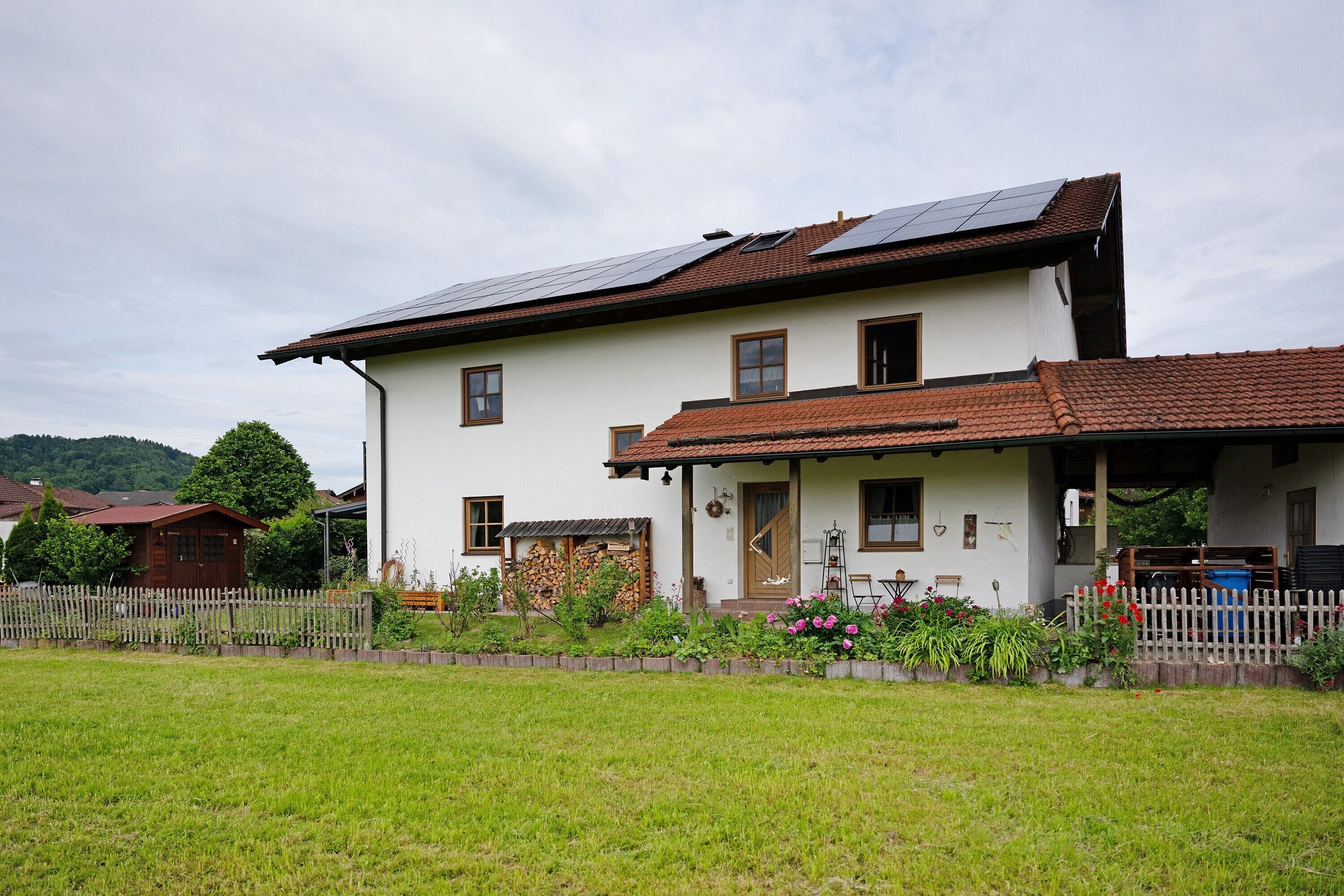 Ein zweistöckiges Haus mit Schrägdach, Solaranlage, Holzzaun und einem Garten mit Blumen und Pflanzen.