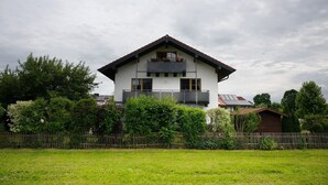 Une maison à deux étages avec un balcon, entourée d’une clôture en bois et de verdure.