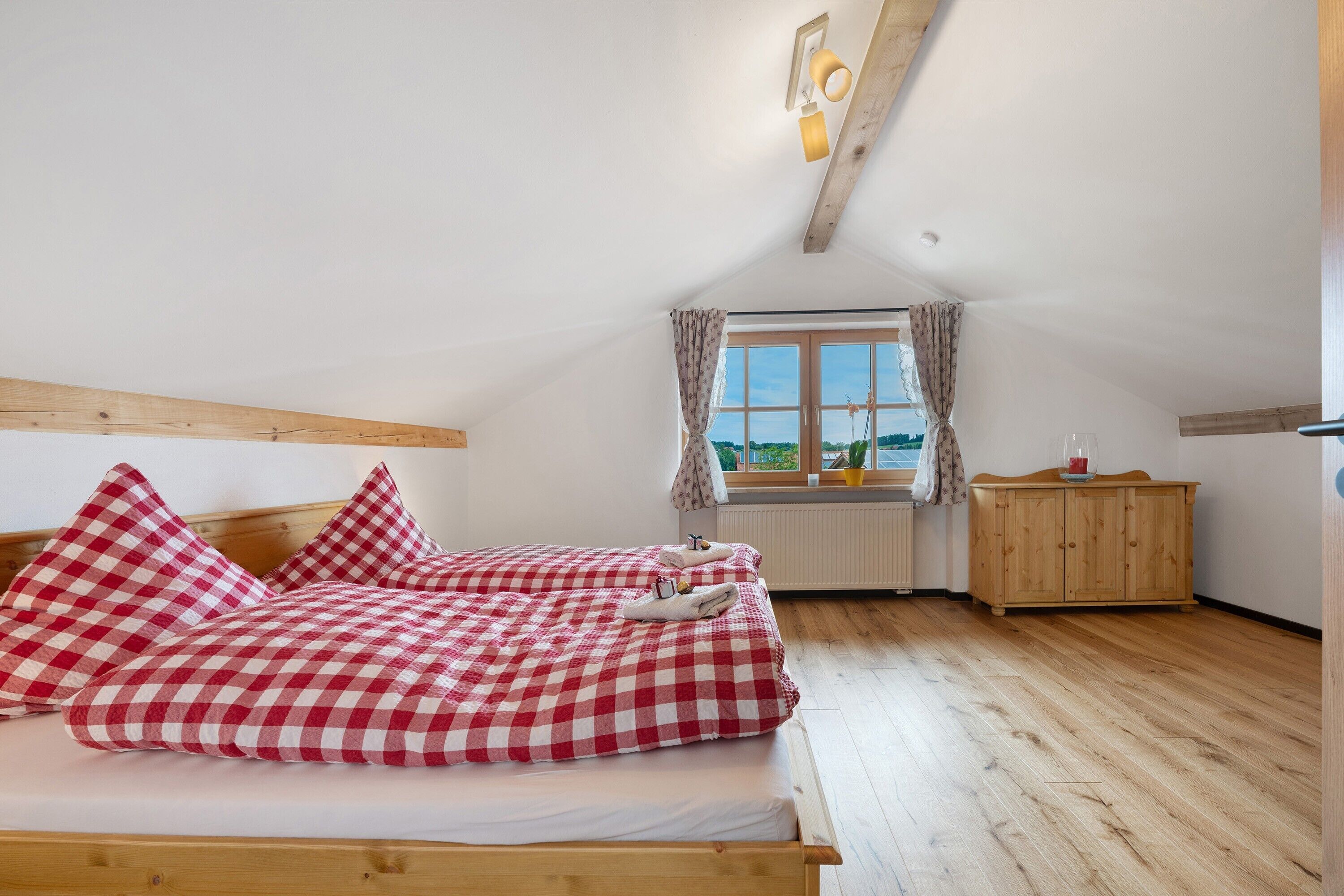 Ein Schlafzimmer mit Holzbalkendecke, einem fenster mit Vorhängen, einem Holzschrank und einem Bett mit Karomuster-Bettwäsche.