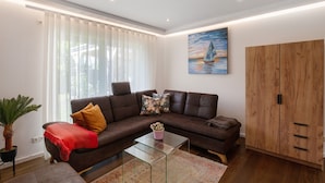 Ein modernes Wohnzimmer mit einer Eckcouch, einem Glastisch, einem Holzschrank und einem großen Fenster mit Jalousien.