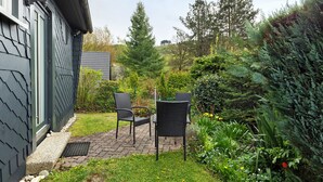 Um pátio com duas cadeiras e uma mesa, rodeado por um jardim com diversas plantas e árvores.