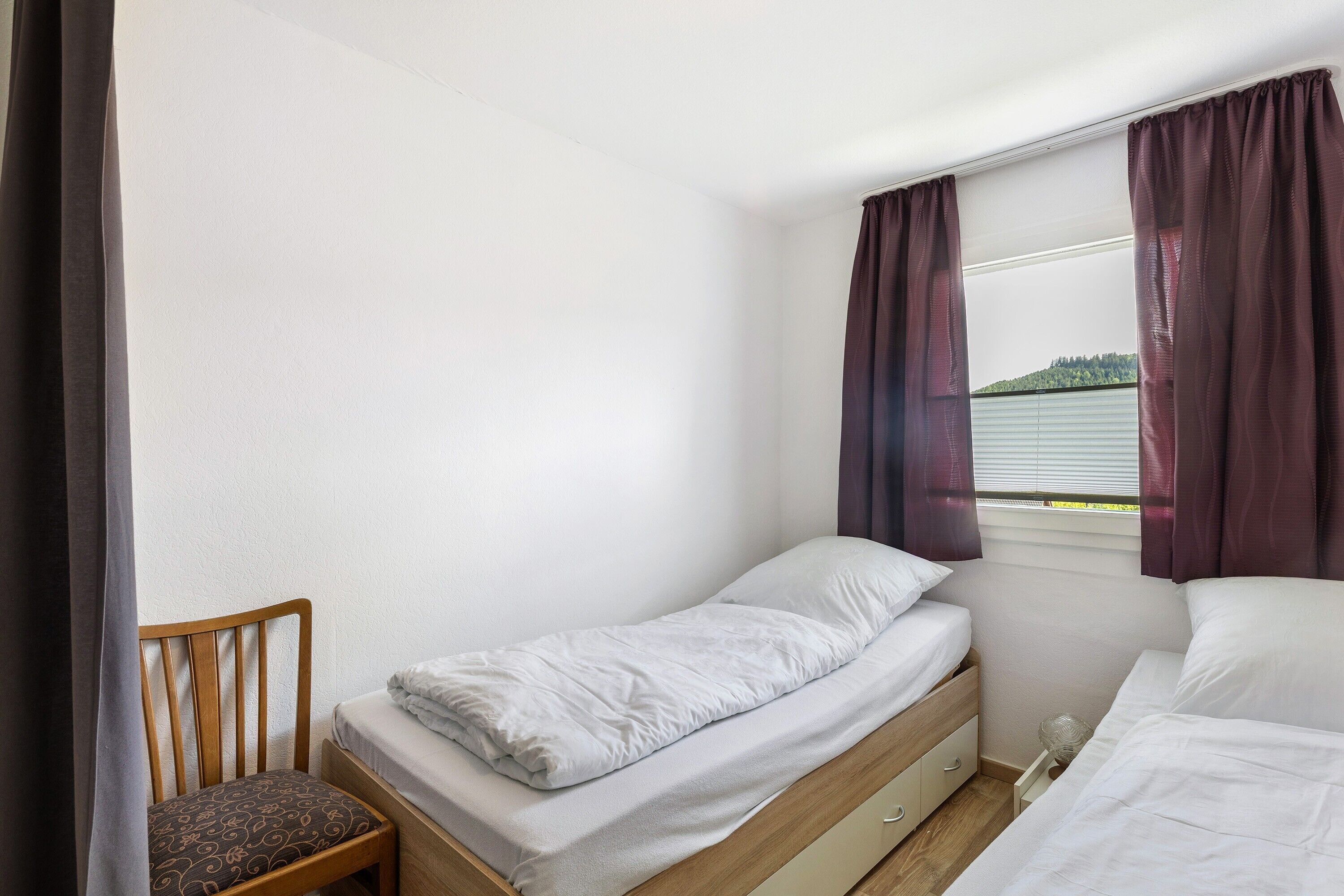 Quarto pequeno com uma cama de solteiro, uma cadeira de madeira, uma janela com cortinas e vista para o verde lá fora.