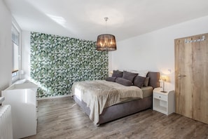 Une chambre à coucher comprenant un lit, des tables de chevet, une armoire en bois, une fenêtre avec des stores et un mur orné d’un motif végétal.