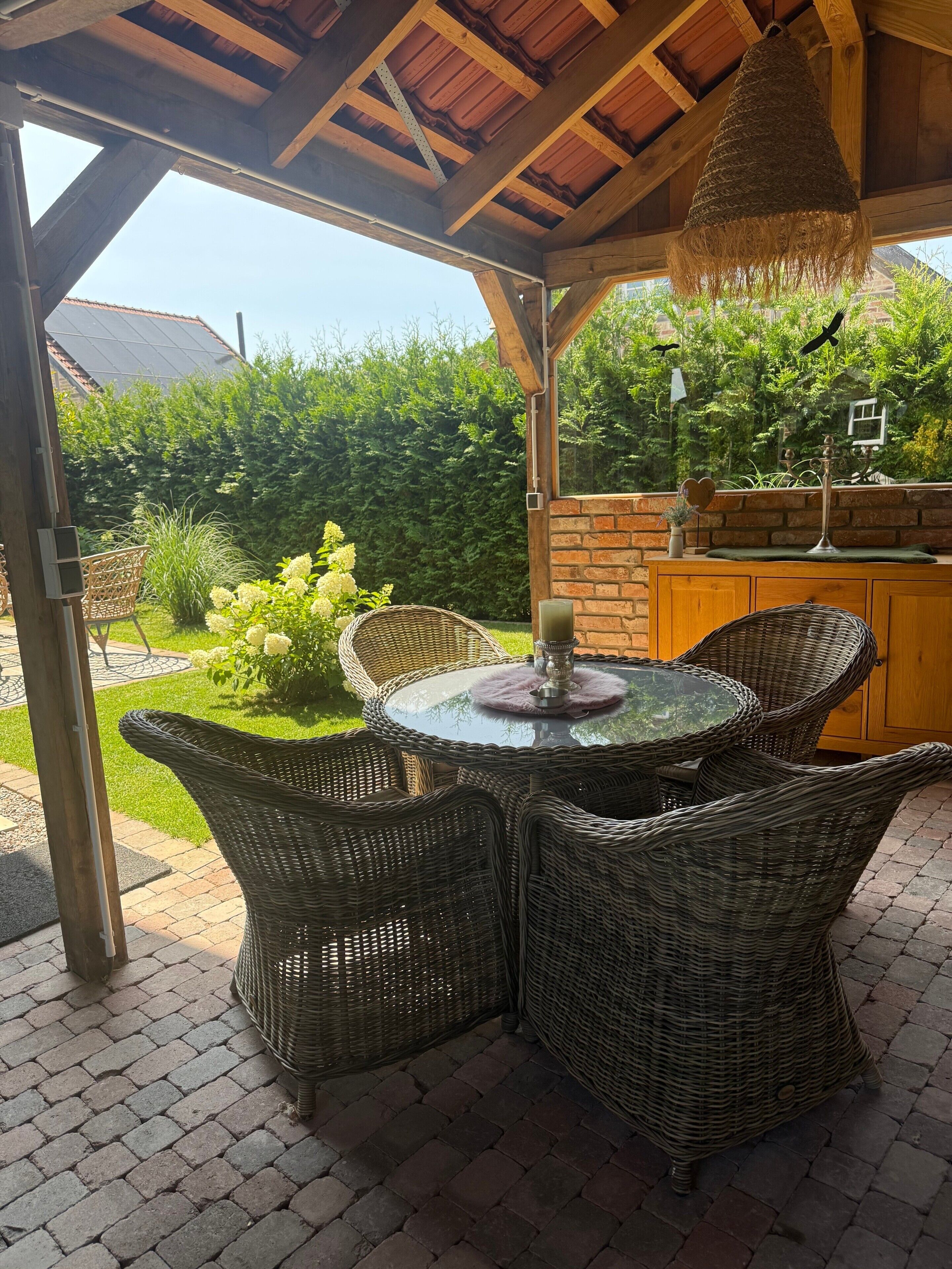 A covered outdoor seating area with wicker furniture, a glass table, and a hanging light fixture.