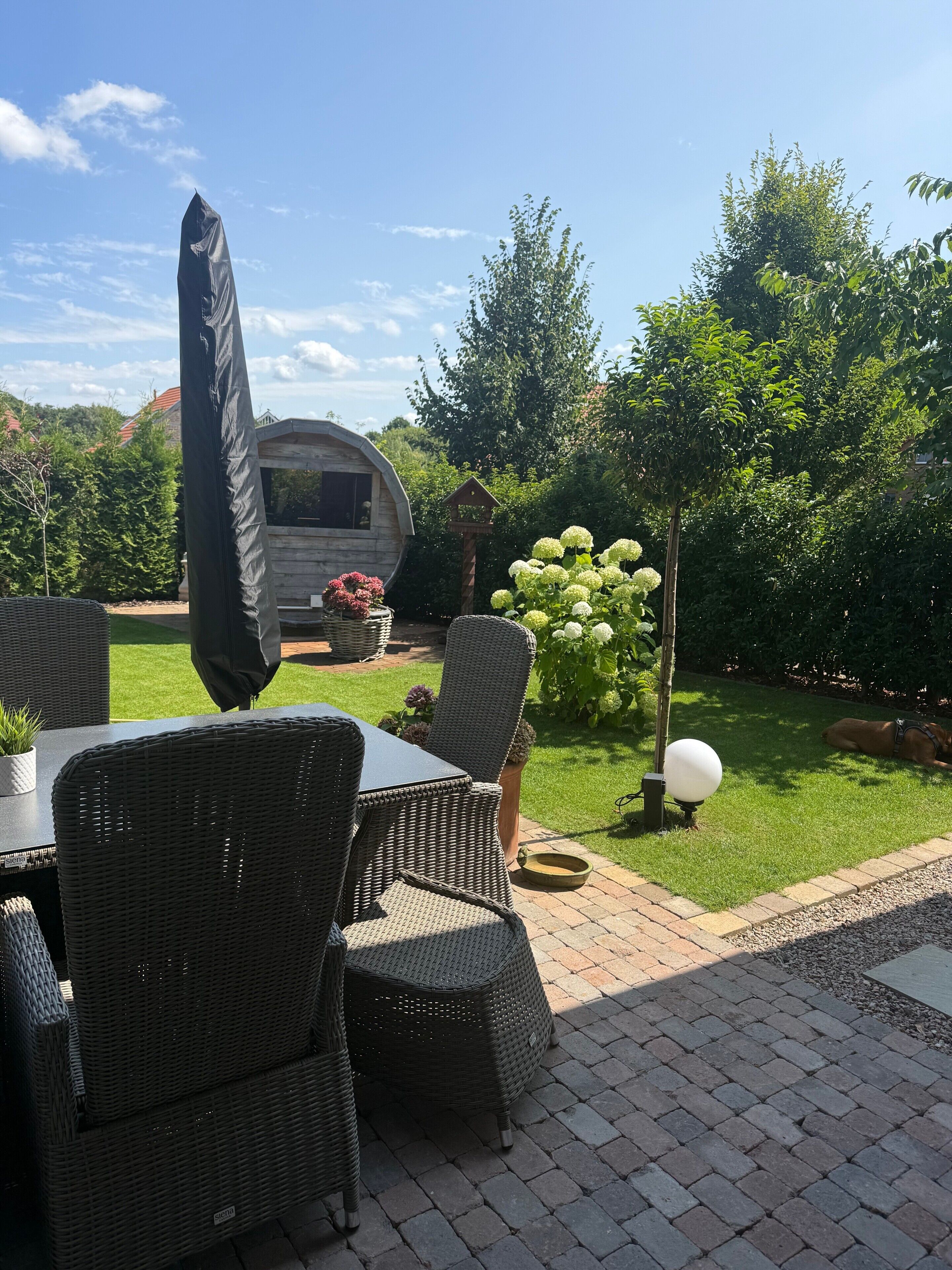 Un espace extérieur aménagé avec des meubles en osier, une table couverte et un jardin comprenant une pelouse et des arbres.