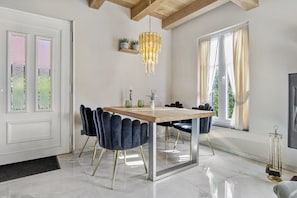 A dining area with a wooden table, dark upholstered chairs, a chandelier, and a large window with curtains.