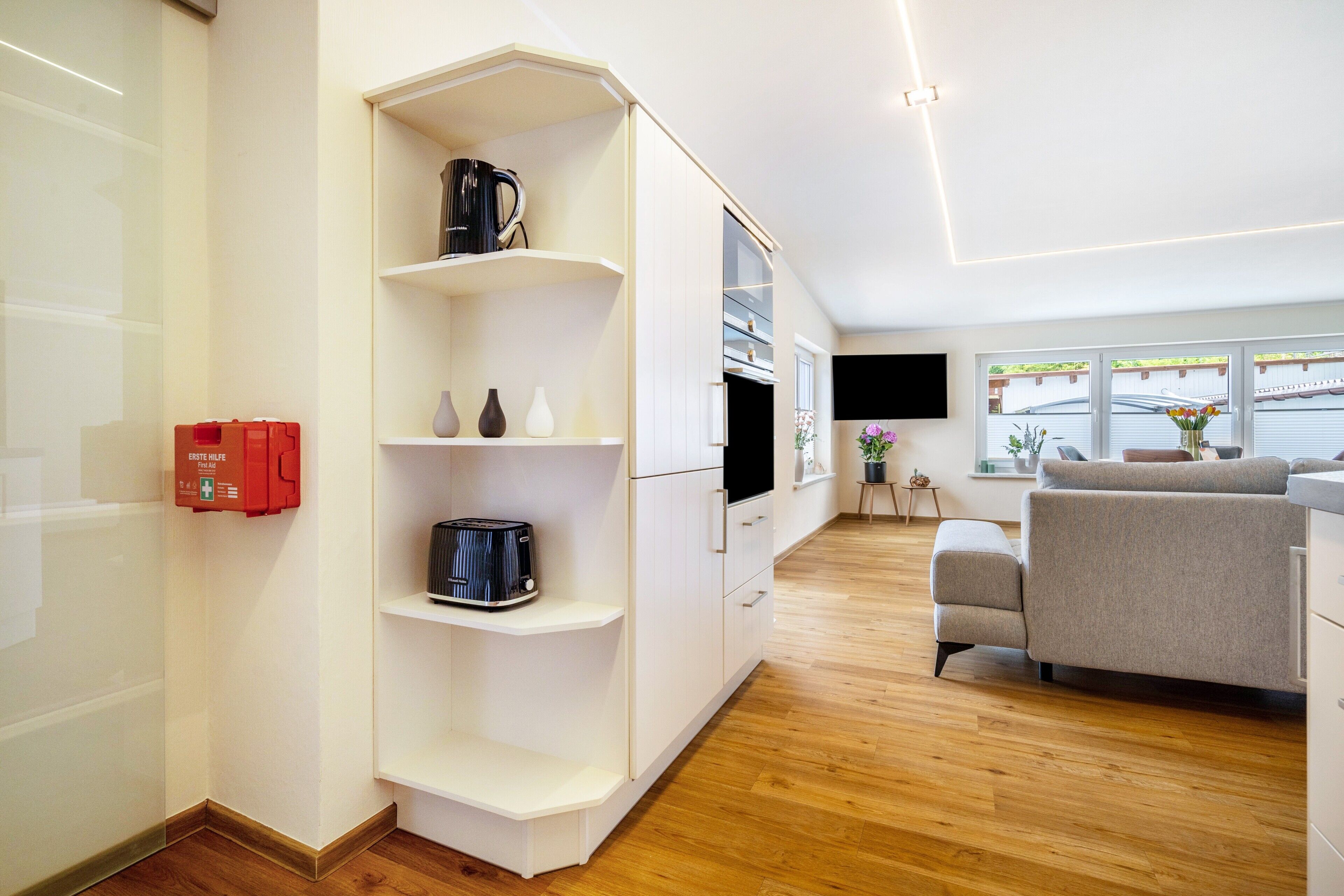 Een moderne woonkamer met een ingebouwde televisie, een bank en een koffiezetapparaat op een wit kastje.