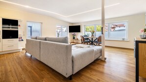 A modern living room with a grey sofa, wooden flooring, a dining area with a table and chairs, and a kitchen with white cabinets and a black oven.