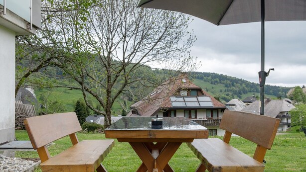 An outdoor seating area with two wooden chairs and a table, a lamp post, and a view of a house and hills in the background.