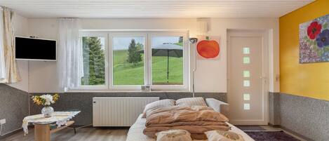 Un dormitorio con cama, una mesita de noche con un jarrón, una ventana con vistas a la vegetación, un televisor montado en la pared y una puerta con interruptor de luz.