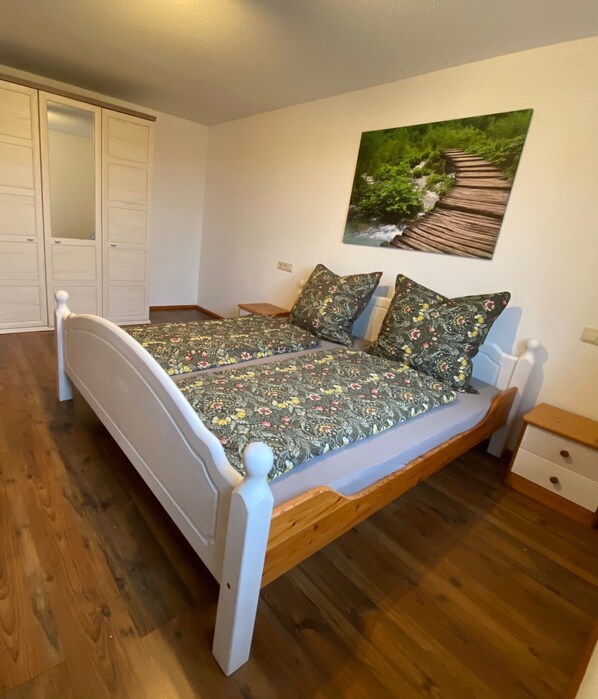 A bedroom with a wooden bed, a white headboard, a wooden nightstand, a small drawer, and a painting on the wall.