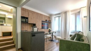 Een moderne keuken met een inbouwmagnetron, een koffieapparaat en een flatscreen-tv waarop 'NETFLIX' wordt weergegeven. Er is een eethoek met een glazen tafel en stoelen, en een groene bank in de woonkamer.