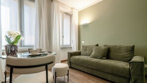 Een woonkamer met een groene bank, een klein rond tafeltje met een bloevas en een raam met gordijnen.