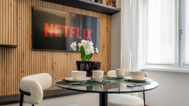 A modern dining area with a glass table, white cushioned chairs, a vase with white flowers, and a wall-mounted TV displaying "NETFLIX".