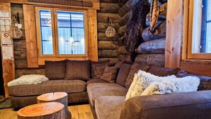 A cozy log cabin interior with a sectional sofa, two wooden stools, and a large window with a view.