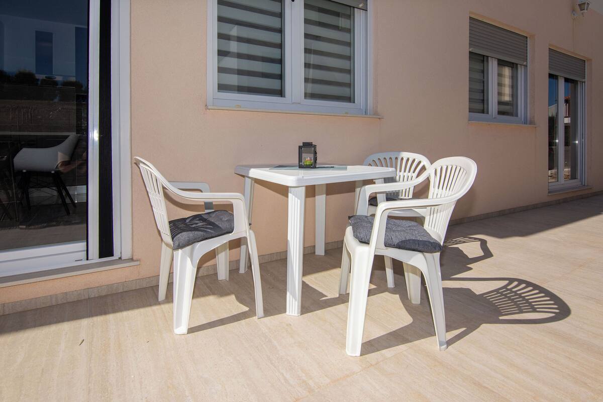 Eine Außenpatio mit einem Tisch und vier weißen Plastikstühlen, einer kleinen Tischlampe und Blick auf ein Gebäude durch eine Glastür.