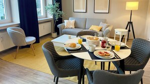 Ein modernes Wohnzimmer mit grauem Sofa, einem Esstisch mit Frühstücksgegenständen, einer Topfpflanze und einem großen Fenster mit Vorhängen.