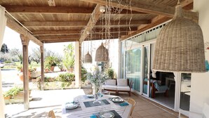 Eine Außenpatio mit einer Holz-Pergola, hängenden Lampen aus Rattangeflecht, einem Esstisch mit Tellern und Gläsern sowie Blick auf einen Garten und ein Gebäude durch Glastüren.